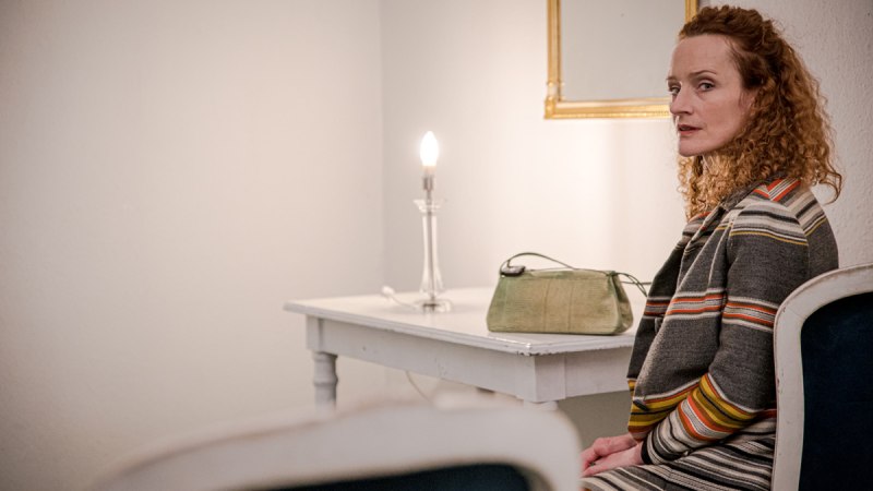 Eine Frau mit lockigem Haar sitzt in einem Raum. Neben ihr ein Tisch mit Lampe und Tasche. Der Raum wirkt schlicht und ruhig., © Thomas Kölsch Eine Frau mit lockigem Haar sitzt in einem Raum. Neben ihr ein Tisch mit Lampe und Tasche. Der Raum wirkt schlicht und ruhig., © Thomas Kölsch