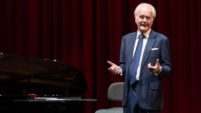 Ein Mann im Anzug spricht auf einer Bühne vor einem roten Vorhang, neben einem Flügel., © Württembergische Staatstheater Stuttgart Ein Mann im Anzug spricht auf einer Bühne vor einem roten Vorhang, neben einem Flügel., © Württembergische Staatstheater Stuttgart