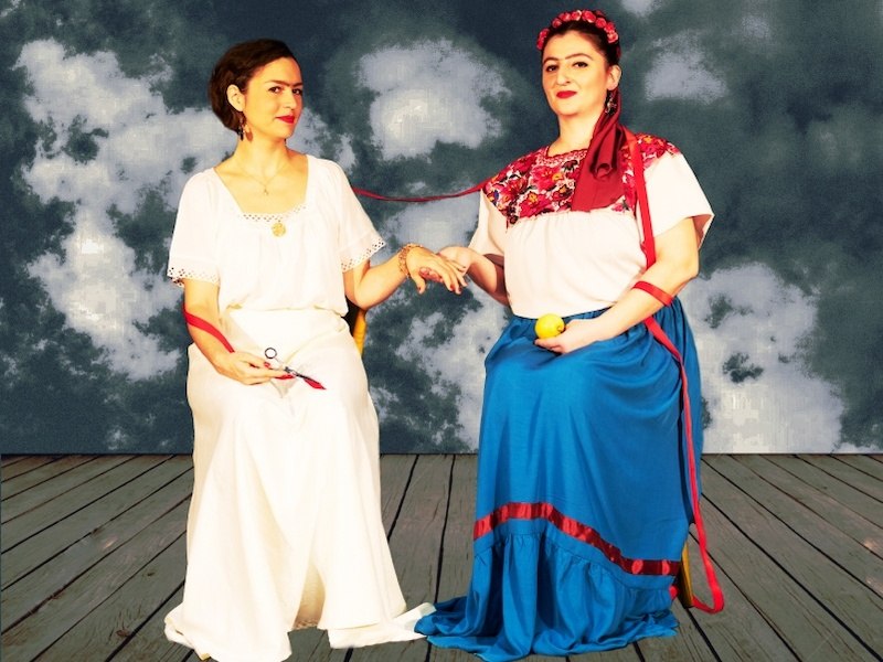 Two women in traditional dress are sitting on a wooden floor. They are connected by a red ribbon. The background shows a cloudy sky., &copy; tri-b&uuml;hne e.V.