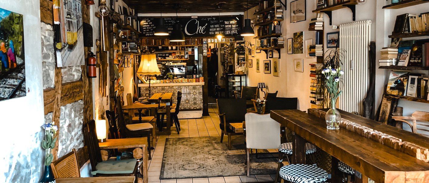 Ein gemütliches Café mit Holzmöbeln, Bücherregalen und Dekorationen an den Wänden. Einladende Atmosphäre mit warmem Licht., © Café Che