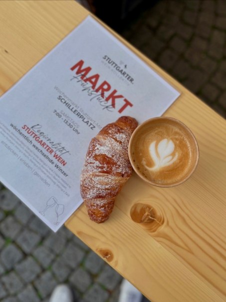 Ein Croissant und ein Cappuccino auf einem Holztisch neben einem Flyer für das Marktfrühstück am Schillerplatz in Stuttgart., © STUTTGARTER.WEIN.KOLLEKTIV! Ein Croissant und ein Cappuccino auf einem Holztisch neben einem Flyer für das Marktfrühstück am Schillerplatz in Stuttgart., © STUTTGARTER.WEIN.KOLLEKTIV!