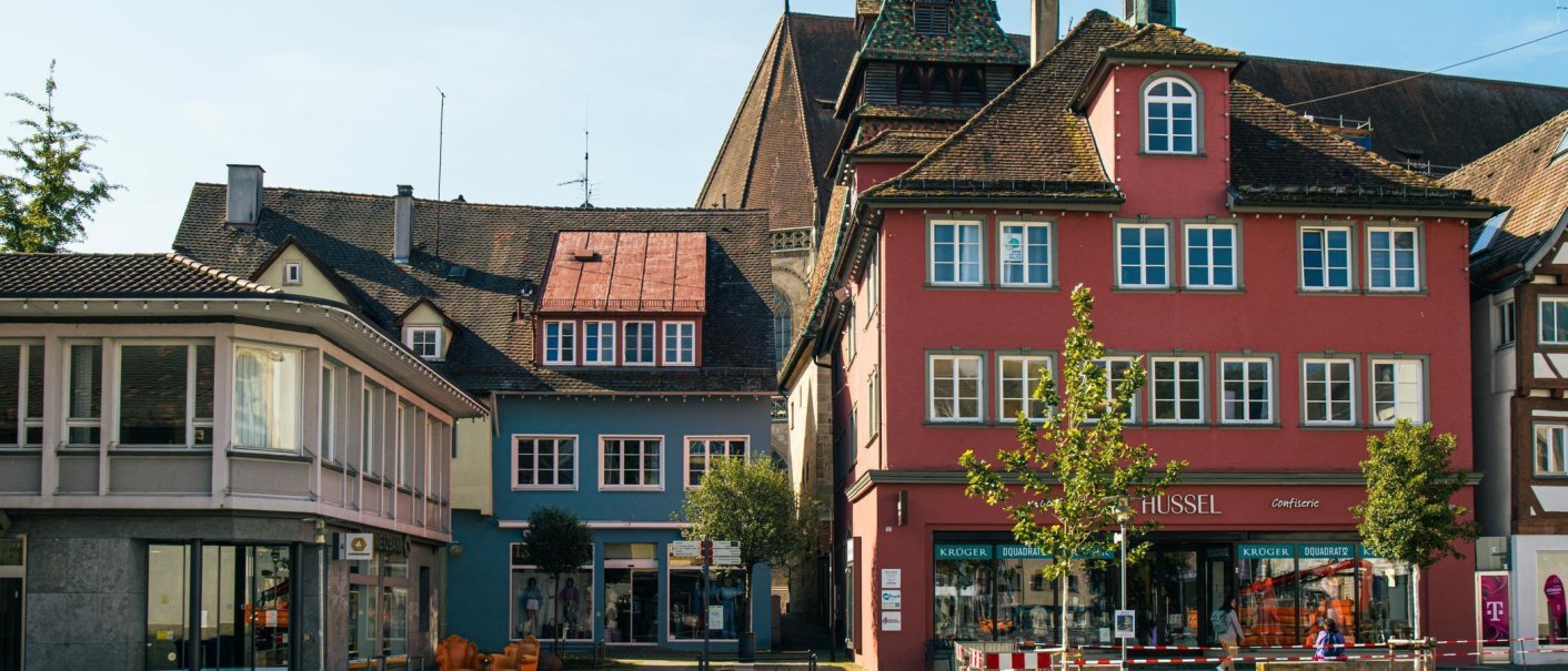 Colorful historic buildings in the old town of Schwäbisch Gmünd, including a red house with stores on the first floor. A tree stands in the foreground., © Stuttgart-Marketing GmbH, Sarah Schmid