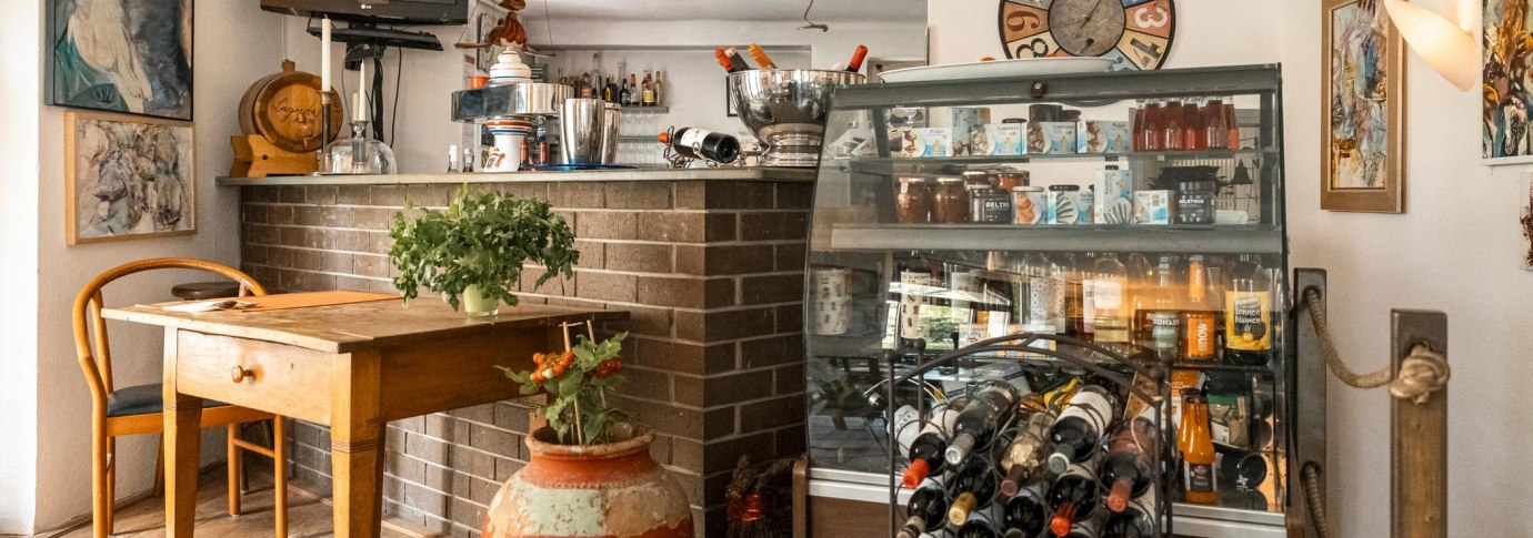 Gemütliches Café mit Holztisch, Weinregal, dekorativen Bildern und Pflanzen. Eine Theke mit Getränken und ein Fernseher sind ebenfalls zu sehen., © SMG, Sarah Schmid