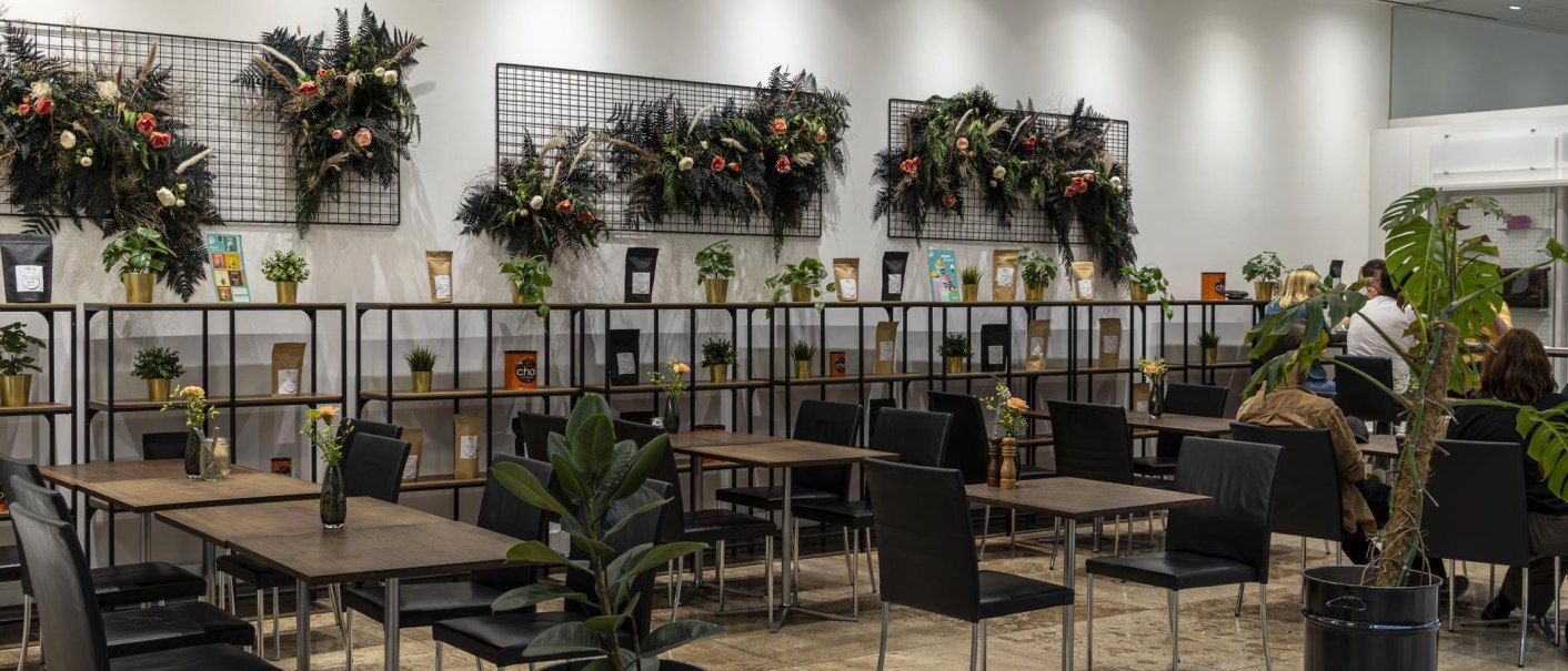 Modernes Café mit Pflanzen und dekorativen Wandarrangements. Schwarze Stühle und Holztische, Regale mit Produkten und Pflanzen., © SMG, Sarah Schmid