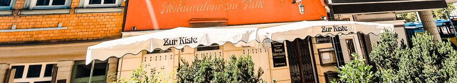 The Zur Kiste wine bar with its red façade, green shutters and awning. Plants adorn the entrance area., © Stuttgart-Marketing GmbH
