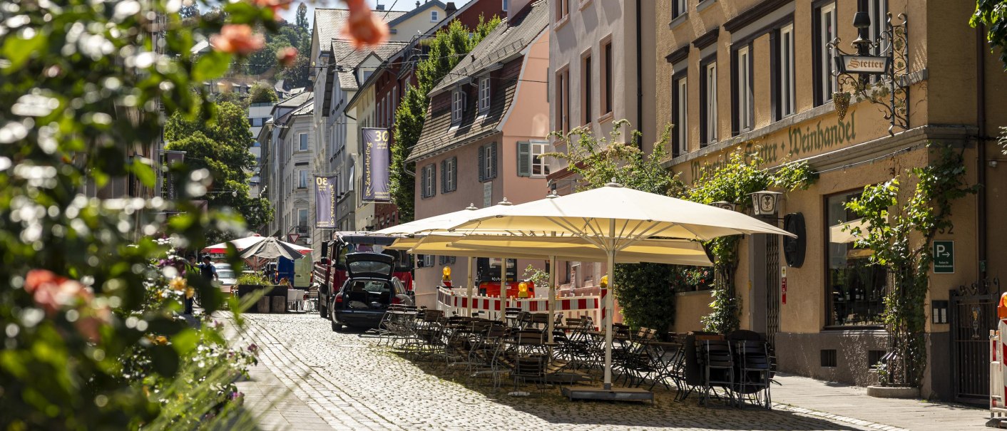 Pflasterstraße im Bohnenviertel mit Weinhaus Stetter, Außensitzbereich unter Sonnenschirmen, umgeben von blühenden Pflanzen und historischen Gebäuden., © Stuttgart Marketing GmbH, Sarah Schmid Pflasterstraße im Bohnenviertel mit Weinhaus Stetter, Außensitzbereich unter Sonnenschirmen, umgeben von blühenden Pflanzen und historischen Gebäuden., © Stuttgart Marketing GmbH, Sarah Schmid