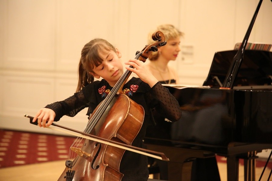 Ein junges Mädchen spielt konzentriert Cello, begleitet von einer Pianistin im Hintergrund. Die Szene findet in einem eleganten Raum statt., © Christian Steeneck Ein junges Mädchen spielt konzentriert Cello, begleitet von einer Pianistin im Hintergrund. Die Szene findet in einem eleganten Raum statt., © Christian Steeneck
