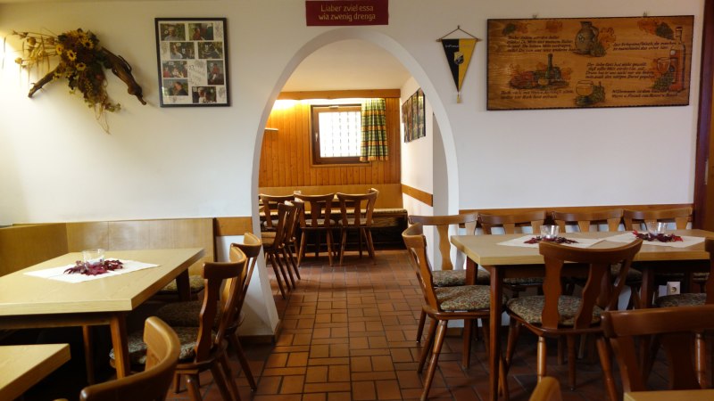 Ein gemütliches Restaurant mit Holztischen und -stühlen, dekoriert mit Bildern und Blumen. Ein Bogen führt in einen weiteren Raum., © Besenwirtschaft Familie Nerz
