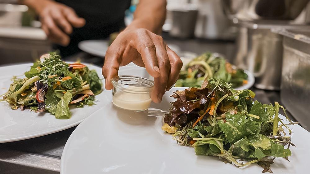 Ein Teller mit frischem Salat wird in einer Küche angerichtet. Eine Hand platziert ein kleines Glas mit Dressing auf dem Teller., © Speisekammer West, Stuttgart