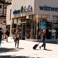 Menschen flanieren vor der Buchhandlung Wittwer-Thalia. Einige sitzen unter Sonnenschirmen in einem Café. Es ist ein sonniger Tag., © Stuttgart Marketing GmbH, Sarah Schmid Menschen flanieren vor der Buchhandlung Wittwer-Thalia. Einige sitzen unter Sonnenschirmen in einem Café. Es ist ein sonniger Tag., © Stuttgart Marketing GmbH, Sarah Schmid