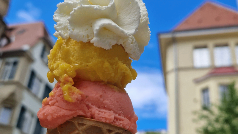 An ice cream cone with three scoops in different colors and whipped cream, against a background of buildings and blue sky., © kesselglueck.de