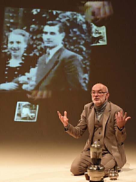 A man sits on a stage and gesticulates. An old photo of a couple is projected in the background., © Theaterhaus Stuttgart e.V. A man sits on a stage and gesticulates. An old photo of a couple is projected in the background., © Theaterhaus Stuttgart e.V.