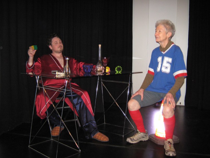 Zwei Personen in einem Raum. Eine Person tr&auml;gt einen roten Bademantel und h&auml;lt einen Zauberw&uuml;rfel. Die andere Person sitzt in Sportkleidung auf einem Verkehrskegel., &copy; Dreigroschentheater