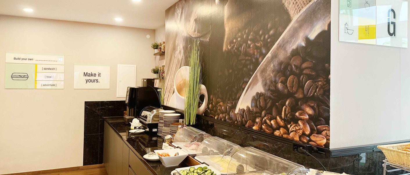 Ein Fr&uuml;hst&uuml;cksbuffet mit Aufschnitt, Gem&uuml;se und Brot. An der Wand ist ein gro&szlig;es Bild von Kaffeebohnen. Schilder ermutigen zur individuellen Gestaltung., &copy; Novum Hospitality