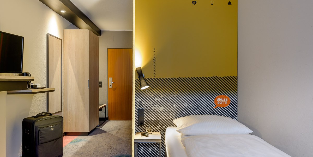 Modernes Hotelzimmer mit Einzelbett, gelber Wand, Schreibtisch, Fernseher und Koffer. An der Wand steht 'Grüß Gott'., © TOMAS