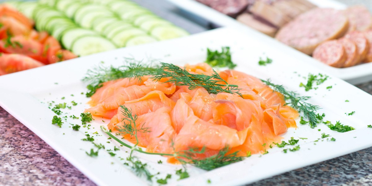 Smoked salmon on a plate, garnished with dill and parsley. In the background are slices of cucumber and tomato, as well as sausage., &copy; ACHAT Hotels Deutschland