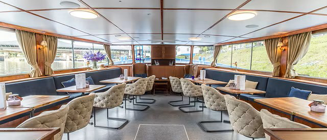 Elegant interior of a ship with tables, upholstered chairs and large windows offering a view of the water., © NeckarFlair MS Wilhelma Betriebsgesellschaft mbH Elegant interior of a ship with tables, upholstered chairs and large windows offering a view of the water., © NeckarFlair MS Wilhelma Betriebsgesellschaft mbH