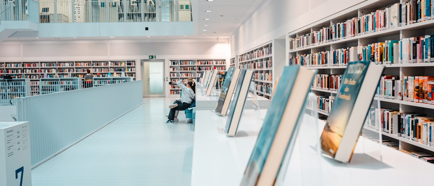Moderne Bibliothek mit weißen Wänden, Bücherregalen und einer Person auf einer Bank. Helle, offene Architektur mit ausgestellten Büchern im Vordergrund., © Stuttgart-Marketing GmbH, Sarah Schmid
