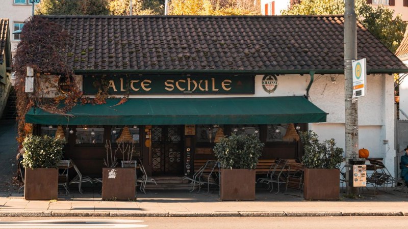 A cozy building with the sign 'Alte Schule' (old school), surrounded by plants. A man sits on a bench while another looks at his cell phone., &copy; SMG Stuttgart Marketing GmbH - Sarah Schmid