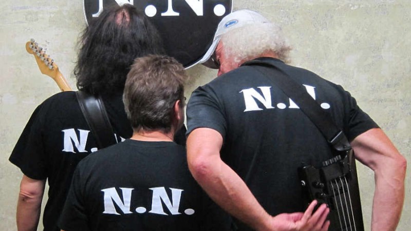 Drei Personen mit schwarzen 'N.N.'-T-Shirts stehen vor einer Trommel mit dem gleichen Logo. Sie halten Instrumente., © NNBros Drei Personen mit schwarzen 'N.N.'-T-Shirts stehen vor einer Trommel mit dem gleichen Logo. Sie halten Instrumente., © NNBros