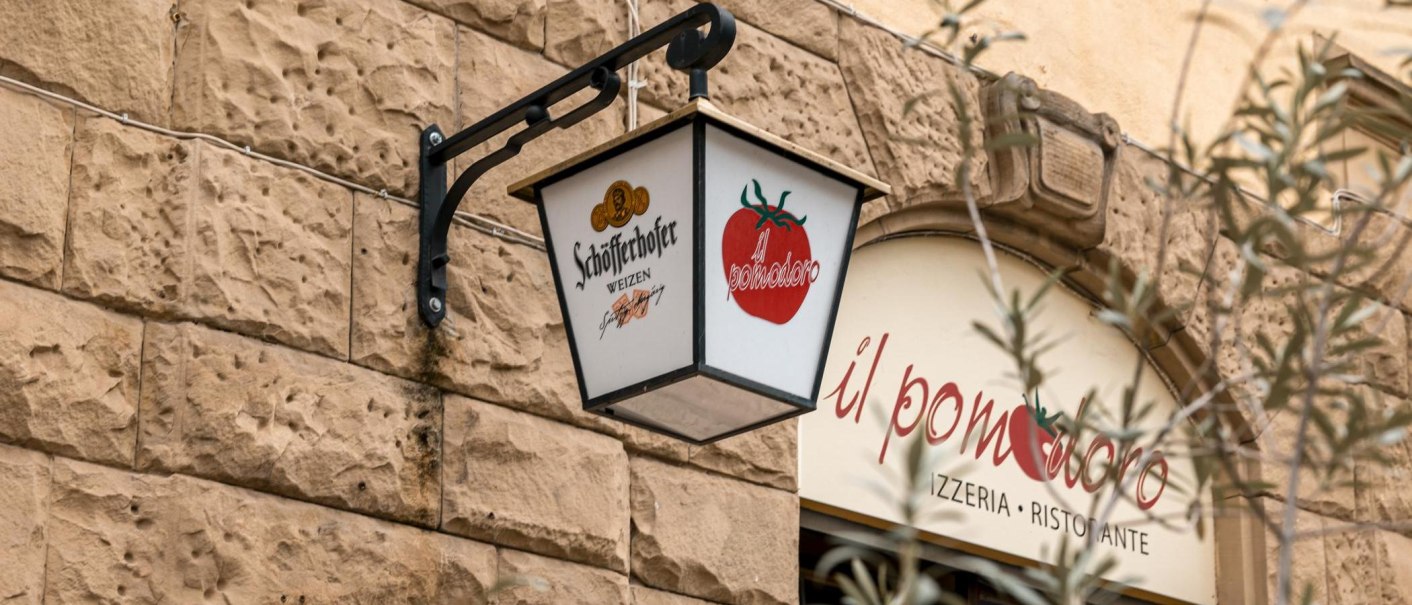 Ein Schild mit Schöfferhofer-Logo und Tomatenmotiv hängt an einer Steinwand vor dem Restaurant 'il pomodoro'., © SMG, Sarah Schmid