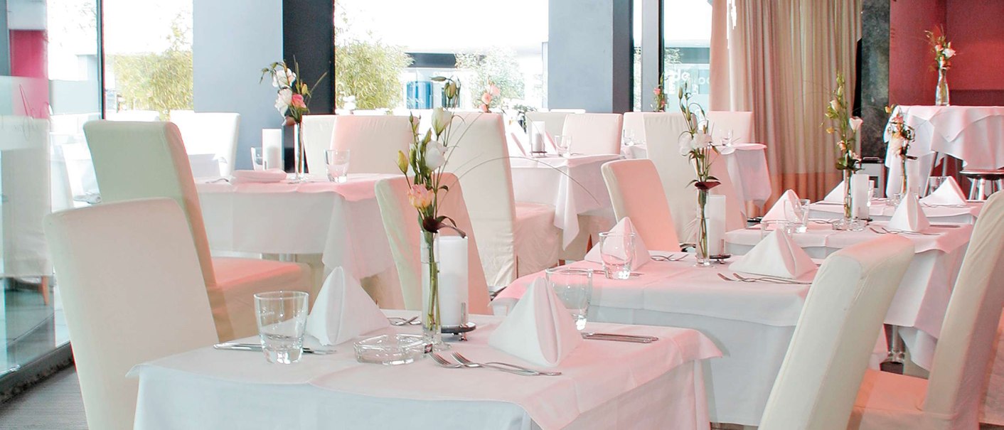 Elegant restaurant with white tablecloths, stylishly laid tables and flower arrangements. Bright, modern furnishings with large windows., © © AMICI Elegant restaurant with white tablecloths, stylishly laid tables and flower arrangements. Bright, modern furnishings with large windows., © © AMICI