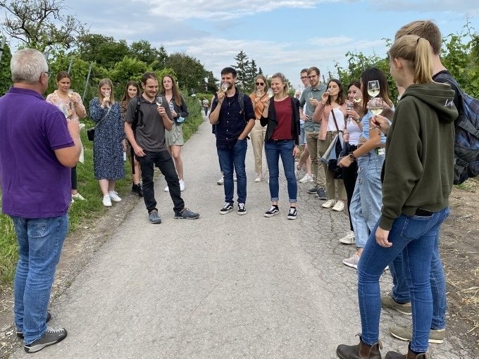 Eine Gruppe von Menschen steht auf einem Weg zwischen Weinreben und genie&szlig;t eine Weinprobe. Sie lachen und unterhalten sich., &copy; Weingenuss Stuttgart