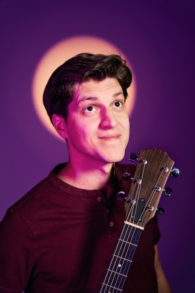A man holds a guitar and looks upwards. The background is purple with a circle of light behind his head, which resembles a halo., &copy; Renitenztheater Stuttgart e.V.