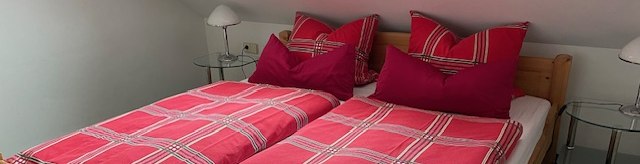 Gemütliches Schlafzimmer mit einem Holzbett, roten karierten Bettbezügen und zwei Nachttischen unter einem Dachfenster., © Weinhof am Württemberg