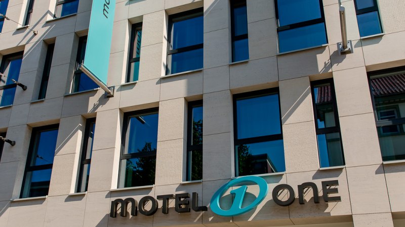 Moderne Fassade eines Geb&auml;udes mit Motel One Logo und blauer Fahne, die im Wind weht. Klare Linien und gro&szlig;e Fenster pr&auml;gen das Design., &copy; Motel One Stuttgart-Bad Cannstatt