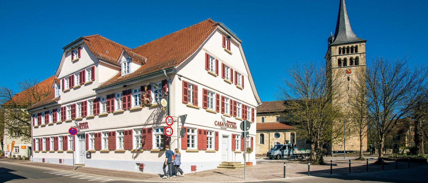 A historic building with red shutters in Sindelfingen, next to a church tower. People walking on the sidewalk, blue sky in the background., © Stuttgart-Marketing GmbH, Sarah Schmid A historic building with red shutters in Sindelfingen, next to a church tower. People walking on the sidewalk, blue sky in the background., © Stuttgart-Marketing GmbH, Sarah Schmid