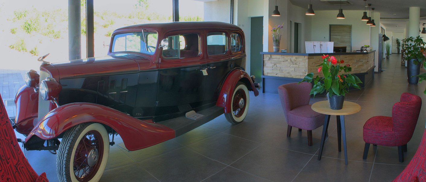 A vintage car in a modern hotel lobby with reception, plants and seating., © TOMAS
