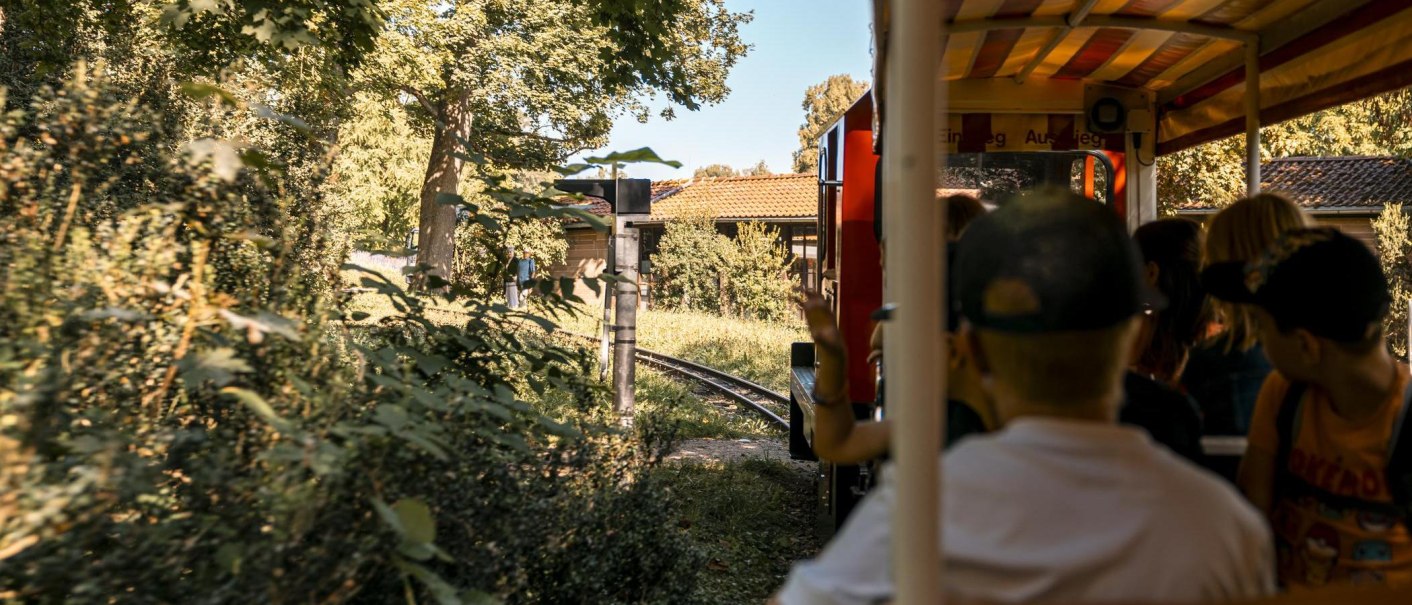 Passagiere in einem offenen Zug, der durch eine grüne Landschaft mit Bäumen und einem Gebäude im Hintergrund fährt., © Stuttgart-Marketing GmbH, Sarah Schmid