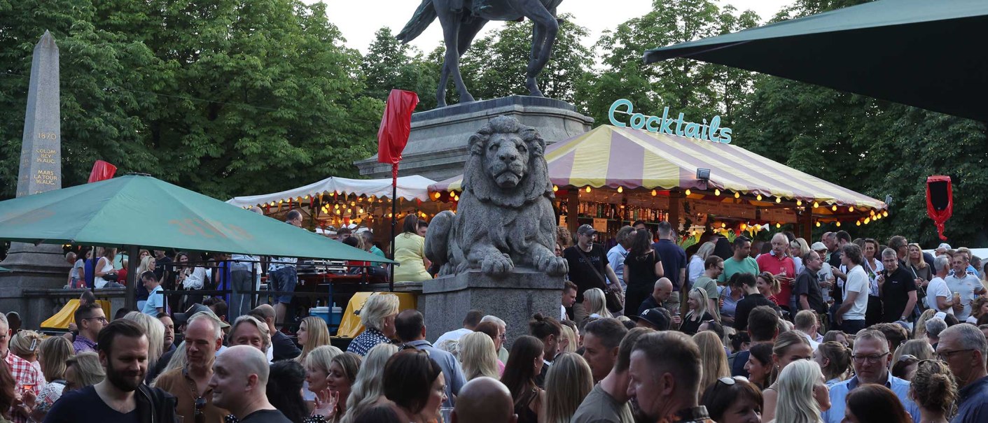 Große Menschenmenge auf einem Markt mit Cocktailstand, Löwenstatue und Bäumen im Hintergrund. Lebhafte Atmosphäre bei Abenddämmerung., © WAGS Große Menschenmenge auf einem Markt mit Cocktailstand, Löwenstatue und Bäumen im Hintergrund. Lebhafte Atmosphäre bei Abenddämmerung., © WAGS