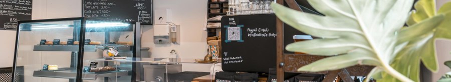A cozy café with a counter, pastry display and drinks menu. Plants decorate the room and a blackboard displays the menu., © SMG, Sarah Schmid