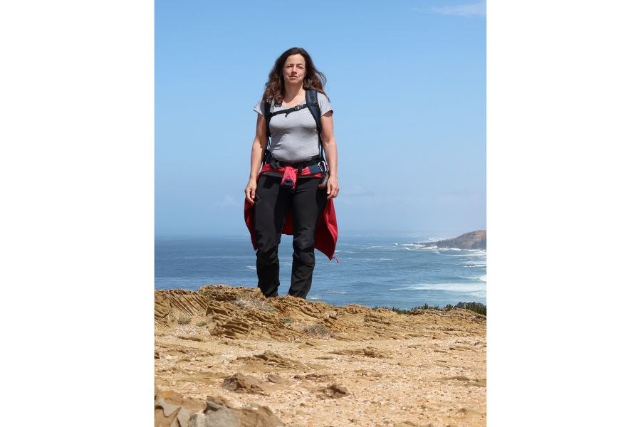 Eine Person steht auf einer felsigen Klippe mit Blick auf das Meer. Sie trägt Outdoor-Kleidung und einen Rucksack. Der Himmel ist blau und klar., © Nina Rühlig