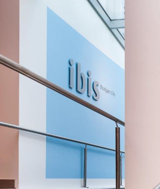 Blick auf das ibis Hotel Stuttgart City Schild in einem modernen Gebäude mit Glasdach und Handlauf., © TOMAS Blick auf das ibis Hotel Stuttgart City Schild in einem modernen Gebäude mit Glasdach und Handlauf., © TOMAS