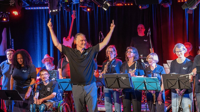 Eine inklusive Band performt auf einer Bühne. Der Dirigent hebt die Arme, während Musiker verschiedener Hintergründe und Fähigkeiten spielen., © Theaterhaus Stuttgart e.V. Eine inklusive Band performt auf einer Bühne. Der Dirigent hebt die Arme, während Musiker verschiedener Hintergründe und Fähigkeiten spielen., © Theaterhaus Stuttgart e.V.