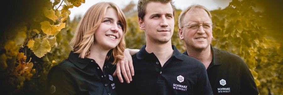 Three people in black clothing with the "Schwarz Weingut" logo stand smiling in a vineyard. They look satisfied and connected., © Weingut Schwarz