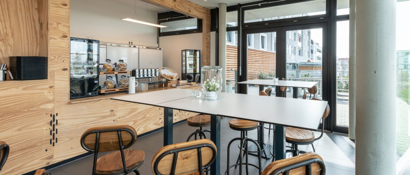 Modern breakfast area with high wooden tables and chairs. Large windows let in plenty of light. A buffet with drinks and pastries in the background., © a2 HOTELS Modern breakfast area with high wooden tables and chairs. Large windows let in plenty of light. A buffet with drinks and pastries in the background., © a2 HOTELS