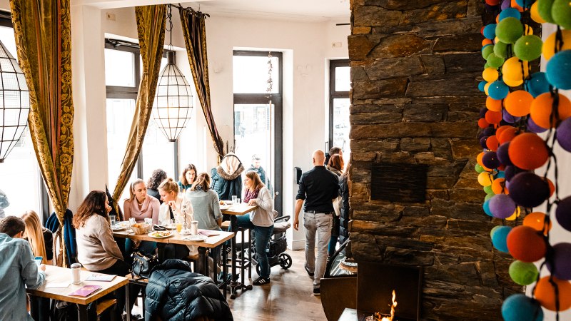 Gemütliches Café mit großen Fenstern, Menschen an Tischen, bunte Kugeln an der Wand und ein Kamin. Warme, einladende Atmosphäre., © Stuttgart-Marketing GmbH, Sarah Schmid