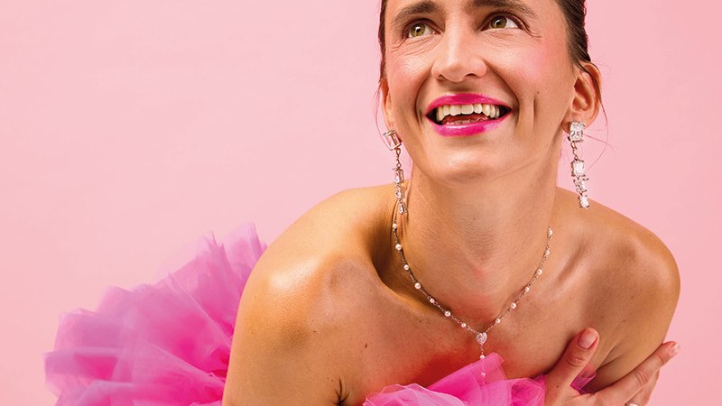 Eine lächelnde Frau in einem rosa Kleid posiert vor einem rosa Hintergrund. Sie trägt auffälligen Schmuck und wirkt fröhlich und elegant., © Renitenztheater Stuttgart e.V. Eine lächelnde Frau in einem rosa Kleid posiert vor einem rosa Hintergrund. Sie trägt auffälligen Schmuck und wirkt fröhlich und elegant., © Renitenztheater Stuttgart e.V.