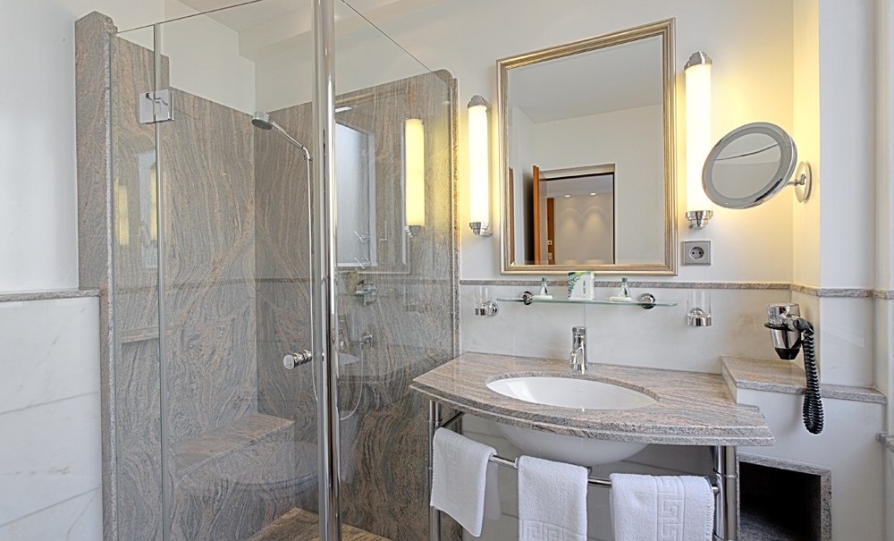 Modernes Badezimmer mit Glasdusche, Waschbecken, großem Spiegel und Handtüchern. Helle Beleuchtung und elegante Marmordetails., © Hotel Royal
