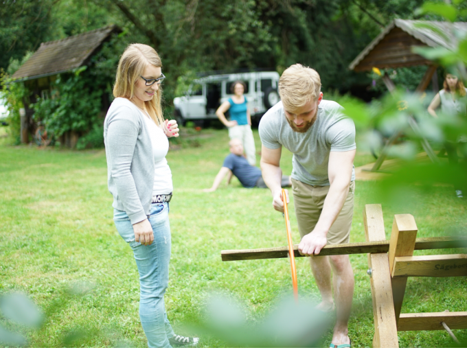 Ein Mann sägt Holz auf einer Wiese, während eine Frau zusieht. Im Hintergrund sind weitere Personen und ein Van zu erkennen., © Cool-Tours StattReisen Ein Mann sägt Holz auf einer Wiese, während eine Frau zusieht. Im Hintergrund sind weitere Personen und ein Van zu erkennen., © Cool-Tours StattReisen