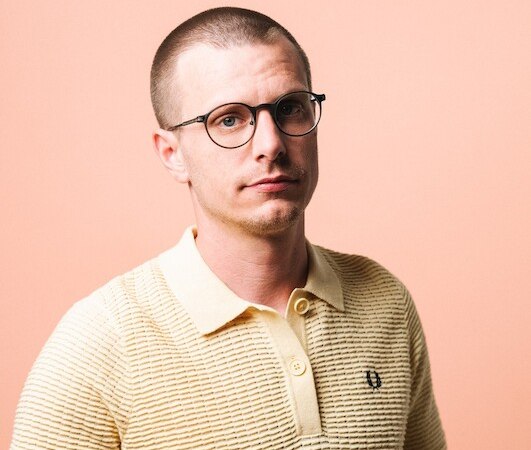 Ein Mann mit Brille und kurzem Haar trägt ein gelbes Polohemd vor einem pfirsichfarbenen Hintergrund., © Rosenau Kultur e.V. Ein Mann mit Brille und kurzem Haar trägt ein gelbes Polohemd vor einem pfirsichfarbenen Hintergrund., © Rosenau Kultur e.V.
