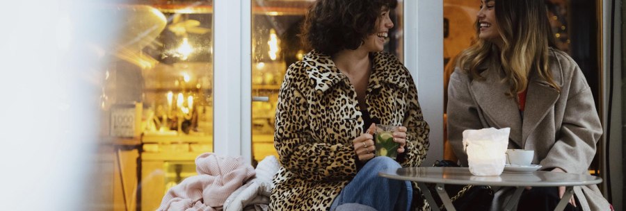 Two women are sitting in a café, laughing and chatting. One is wearing a leopard coat, the other a gray coat. There are drinks on the table., © © Stuttgart-Marketing GmbH, Alwin Maigler