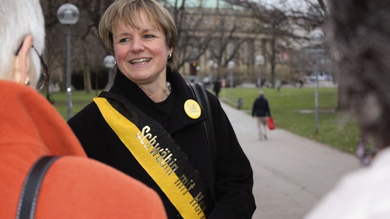 Eine Frau mit einer Schärpe 'Schwäbin mit Herz' lächelt und spricht mit anderen Personen in einem Park., © Stuttgart-Marketing GmbH