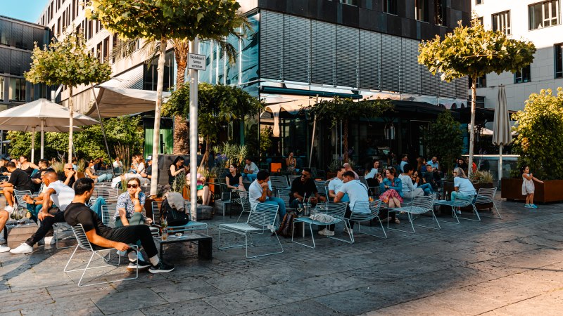 Menschen genießen das sonnige Wetter in einem belebten Außenbereich eines Cafés. Moderne Gebäude und Bäume umgeben die Szene., © SMG, Sarah Schmid