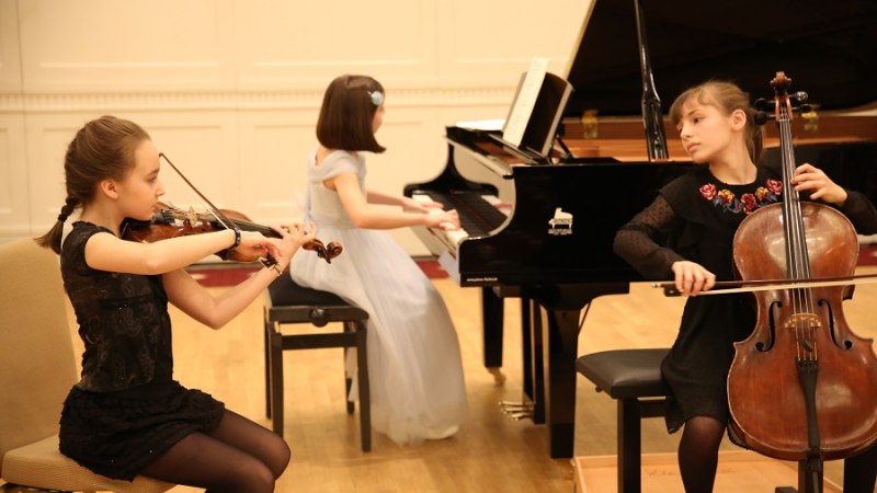 Drei Mädchen spielen Violine, Klavier und Cello in einem Konzertsaal. Sie tragen elegante Kleider und konzentrieren sich auf ihre Instrumente., © Christian Steeneck