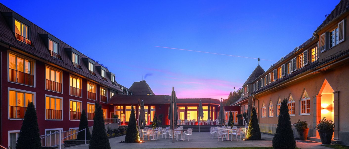 The Waldhotel Stuttgart at dusk, with illuminated windows and a fireplace in the garden. The sky is blue with a pink stripe., &copy; Waldhotel Stuttgart GmbH